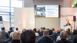 Ein Mann steht an einem Podium des LOPEC Forums in einer Halle und hält einen Vortrag vor einem interessierten Publikum.