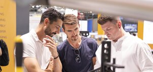 Drei Männer diskutieren an einem Messestand der automatica über industrielle Lösungen; sie begutachten gemeinsam technische Unterlagen und Exponate.