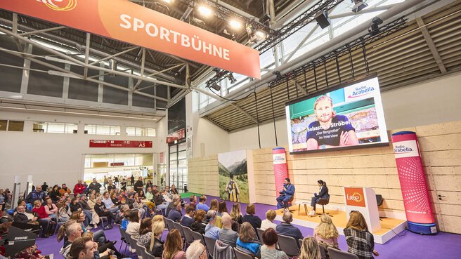 Interview auf der „Sportbühne“ der Messe f.re.e mit großem Publikum, einem Moderator und Sebastian Ströbl als Gast, während ein Bildschirm den Gast zeigt.