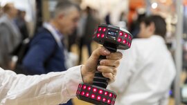 Nahaufnahme einer Hand, die eine Hantel mit elektrischen Komponenten enthält. Im unscharfen Hintergrund unterhalten sich mehrere Messebesucher.