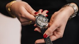 A black and silver watch with visible cogs is held by a pair of women’s hands with red-polished nails, adorned with a watch, bangles and rings on their fingers.