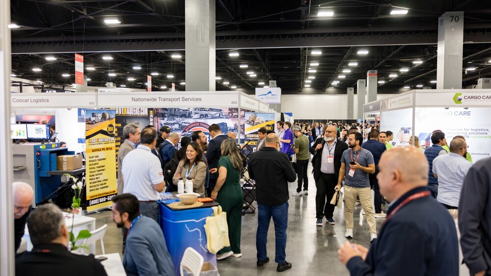 A view of a busy exhibition hall at transport logistic Americas, featuring numerous exhibition stands with advertising banners and numbered concrete pillars in between.