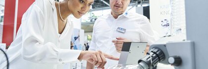 Eine Frau in einer weißen langen Bluse zeigt auf ein wissenschaftliches Gerät, während ein Mann in Business-Kleidung daneben steht. Beide sind auf einem Messestand.