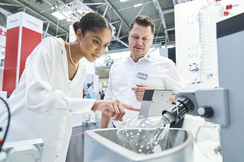 Eine Frau in einer weißen langen Bluse zeigt auf ein wissenschaftliches Gerät, während ein Mann in Business-Kleidung daneben steht. Beide sind auf einem Messestand.