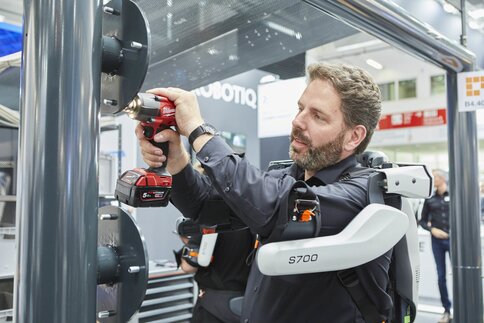 A man uses a cordless screwdriver on a metal structure at an exhibition stand, supported by an exoskeleton.