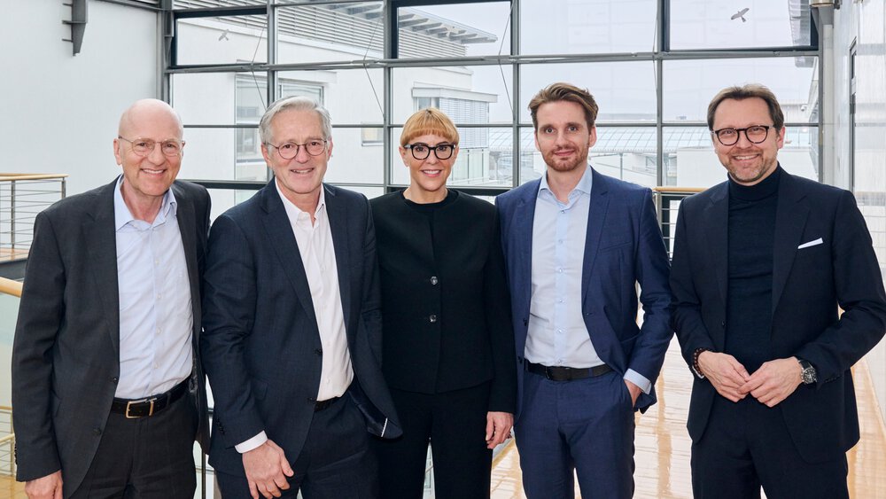  Fünf Personen in Geschäftskleidung stehen lächelnd in einem modernen Büro mit Glaswänden.