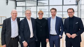  Fünf Personen in Geschäftskleidung stehen lächelnd in einem modernen Büro mit Glaswänden.