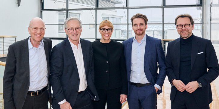  Fünf Personen in Geschäftskleidung stehen lächelnd in einem modernen Büro mit Glaswänden.