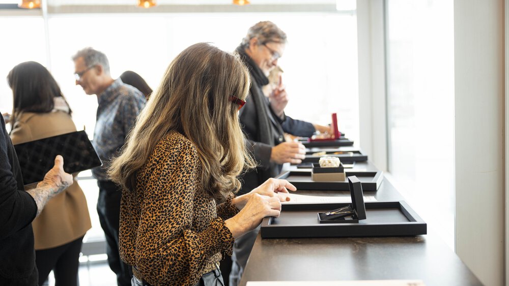 People look at pieces of jewellery on tables with backlighting and large windows.