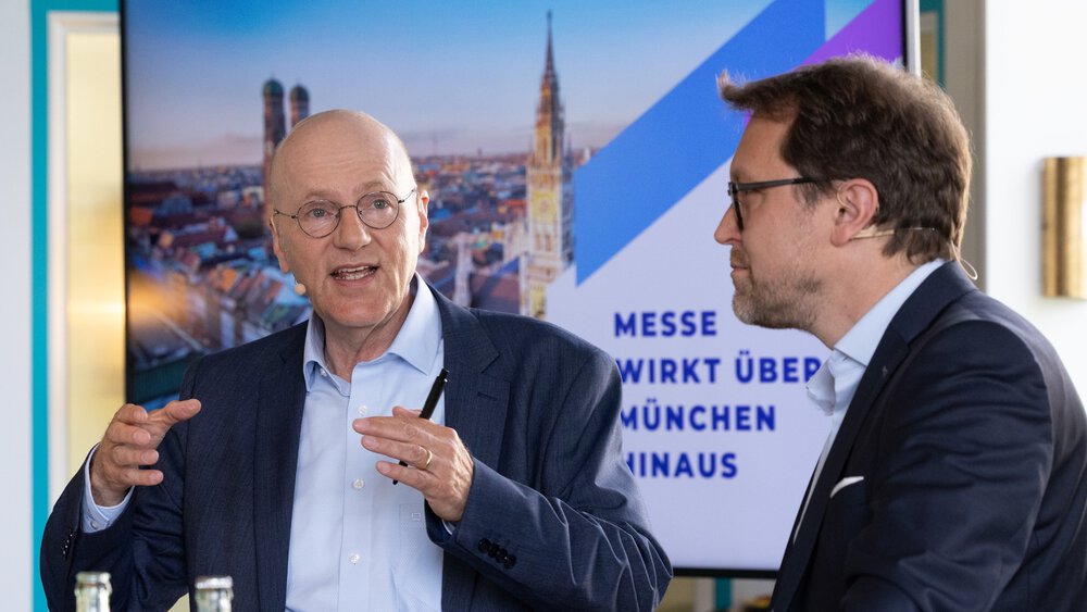 Two men in suits—one bald and gesticulating, the other with dark hair and a beard—are standing in front of a screen showing a presentation.
