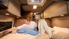 Zwei lachende Frauen in einem Wohnmobil mit Holzvertäfelung, eine sitzt auf dem Bett, während die andere versucht ein Fenster zu öffnen.