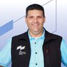 A smiling man in a blue shirt and a black waistcoat is standing in front of a geometric blue and white background.