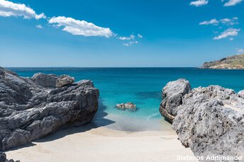 Ein felsiger Strand mit klarem, blauem Wasser und hellem Himmel, kleine Wellen, die an den Sandstrand plätschern.