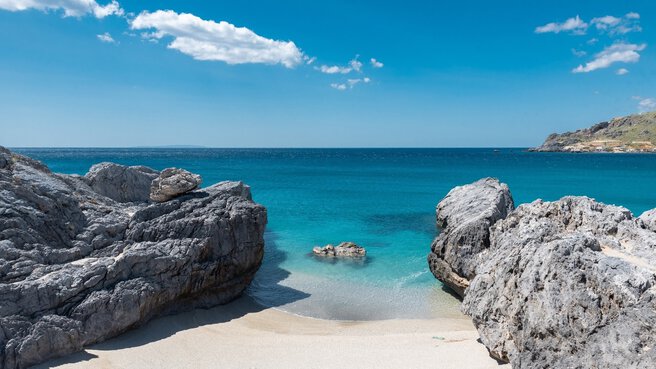 Ein felsiger Strand mit klarem, blauem Wasser und hellem Himmel, kleine Wellen, die an den Sandstrand plätschern.
