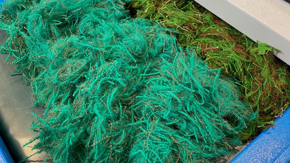 Haufen aus recycelten Textilfasern in Türkis und Grün liegen nebeneinander auf einer industriellen Arbeitsfläche.