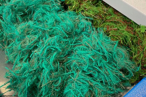 Piles of recycled textile fibers in turquoise and green lying next to each other on an industrial work surface.