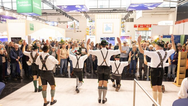 Several men in Bavarian traditional dress perform a traditional dance on a stage, while numerous trade fair visitors watch the performance.