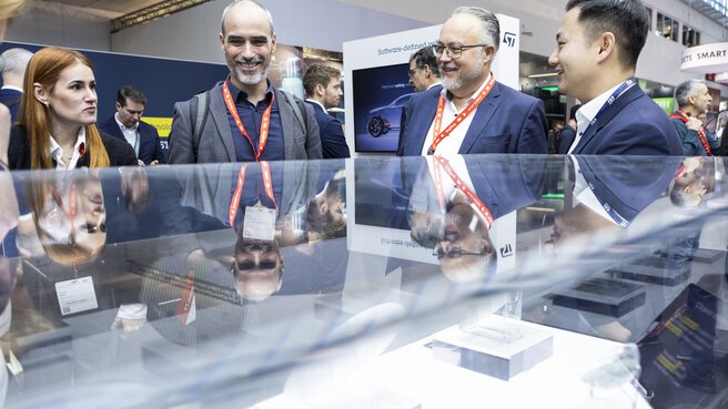 Vier Personen in Geschäftskleidung unterhalten sich und lächeln am Stand einer Messe oder Konferenz.