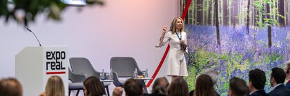 A woman is speaking on a stage at EXPO REAL. She stands next to chairs and a podium, against the backdrop of a forest.
