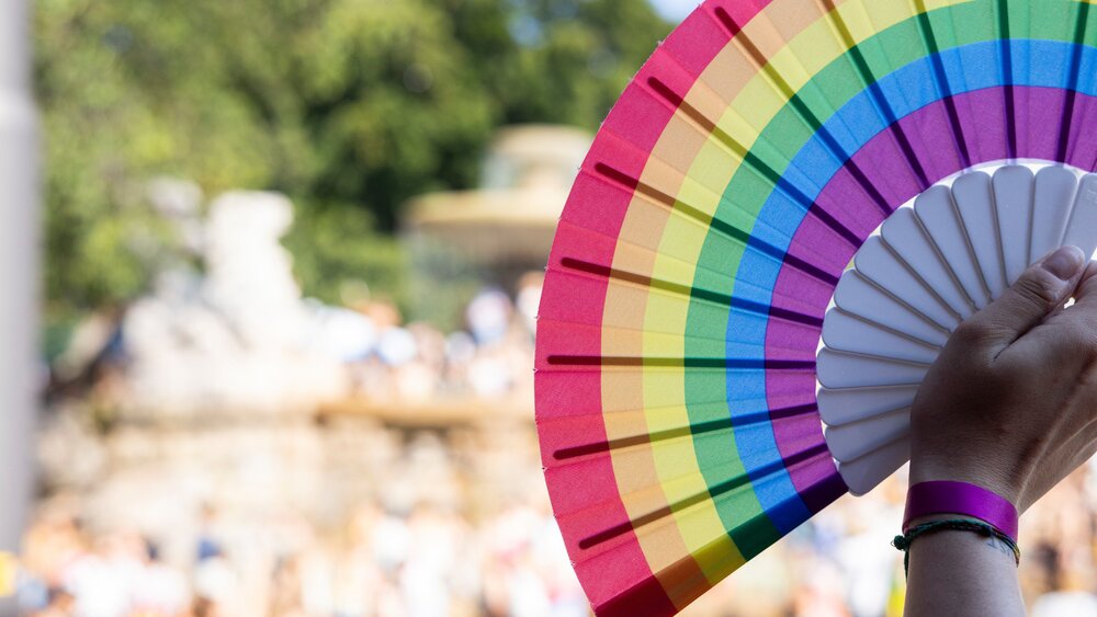 Outdoors, a hand is holding a rainbow fan; in the background, a crowd of people and trees can be seen blurred.