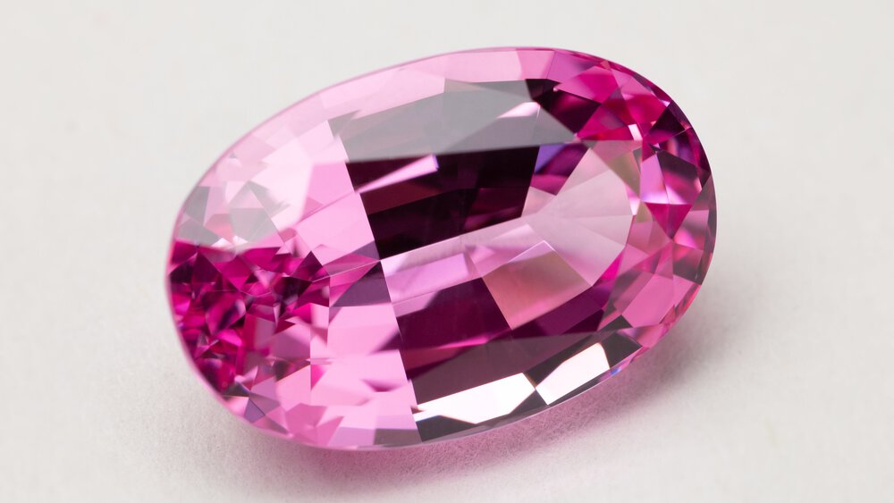 Close-up view of a pink gemstone with multiple, even cuts in an oval shape, resting on a white base.