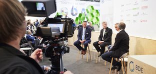 View over the shoulder of a cameraman of a panel discussion with 4 participants on the BDE stage in an exhibition hall.
