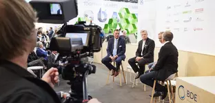 View over the shoulder of a cameraman of a panel discussion with 4 participants on the BDE stage in an exhibition hall.