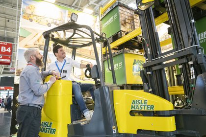 An exhibitor sits grinning in the driver's seat of a yellow forklift truck, a visitor leans against the machine and talks to him.