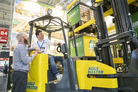 An exhibitor sits grinning in the driver's seat of a yellow forklift truck, a visitor leans against the machine and talks to him.