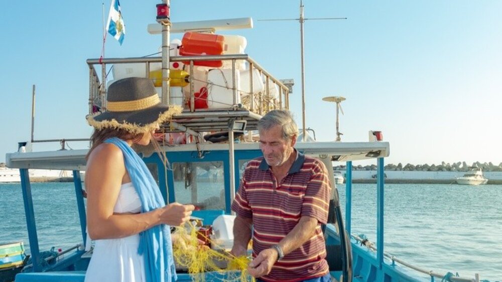 Frau und Mann stehen auf einem blauen Fischerboot und entwirren gelbe Fischernetze unter einem klaren Himmel.