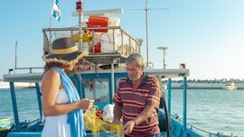 Frau und Mann stehen auf einem blauen Fischerboot und entwirren gelbe Fischernetze unter einem klaren Himmel.