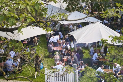 Blick von oben durch Baumzweige auf einen Biergartenbereich mit Sonnenschirmen, Tischen, Stühlen und zahlreichen Messebesuchern, die Pause machen.