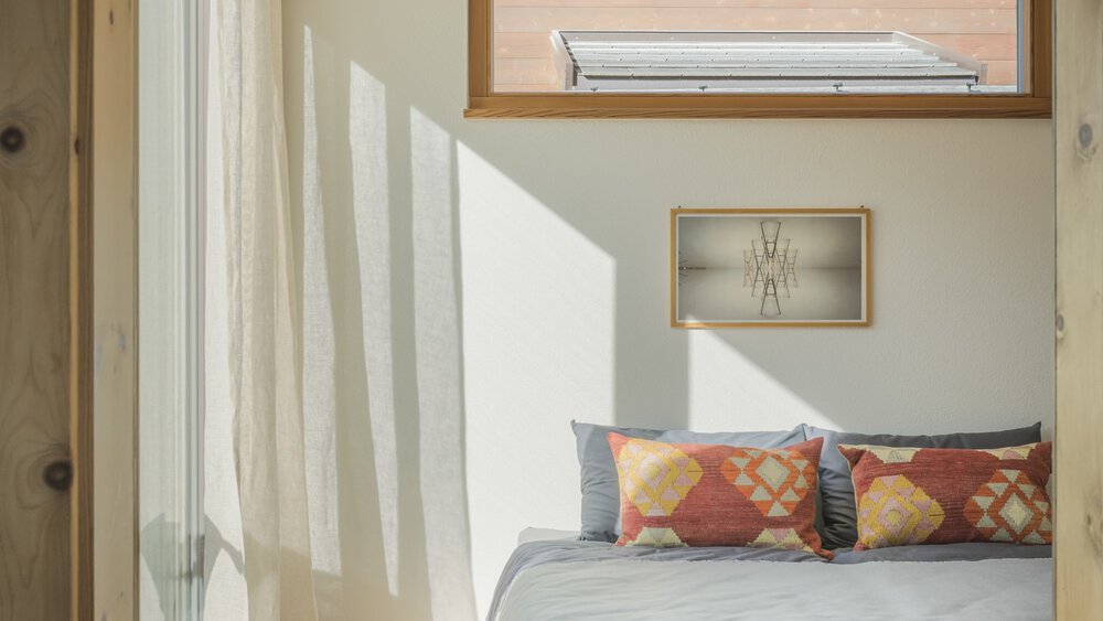 Sonnendurchflutete Zimmerecke mit weißen Vorhängen, einem kleinen Gemälde und Kissen auf einer Bank, die sanfte Schatten werfen.