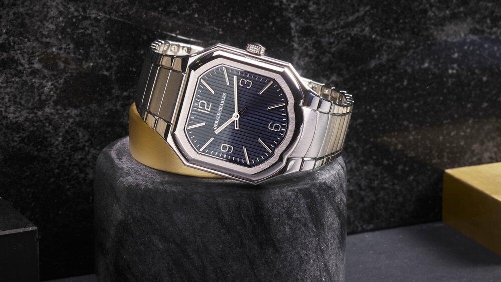 Silver clock by Gerald Charles with hexagonal and black dial on a stone stand against a dark textured background.