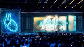 Ein großes Publikum blickt auf eine futuristische Bühne mit einem holografischen Herz und dem Text „Ethical Commitment“, während die Menschen das Ereignis mit ihren Handys aufnehmen.