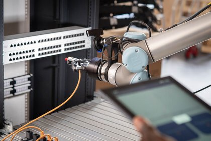 A robotic arm is plugging a network cable into a patch panel. A person in the foreground is holding a tablet.