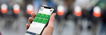 One hand holds a smartphone on which the interface of the IFAT Munich app with green navigation buttons is displayed.