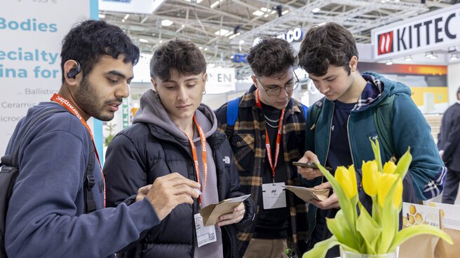 Vier junge Leute stehen an einem Messestand zusammen und betrachten ausgestellte Papiertüten auf denen Honigwaben und eine Biene aufgedruckt sind.