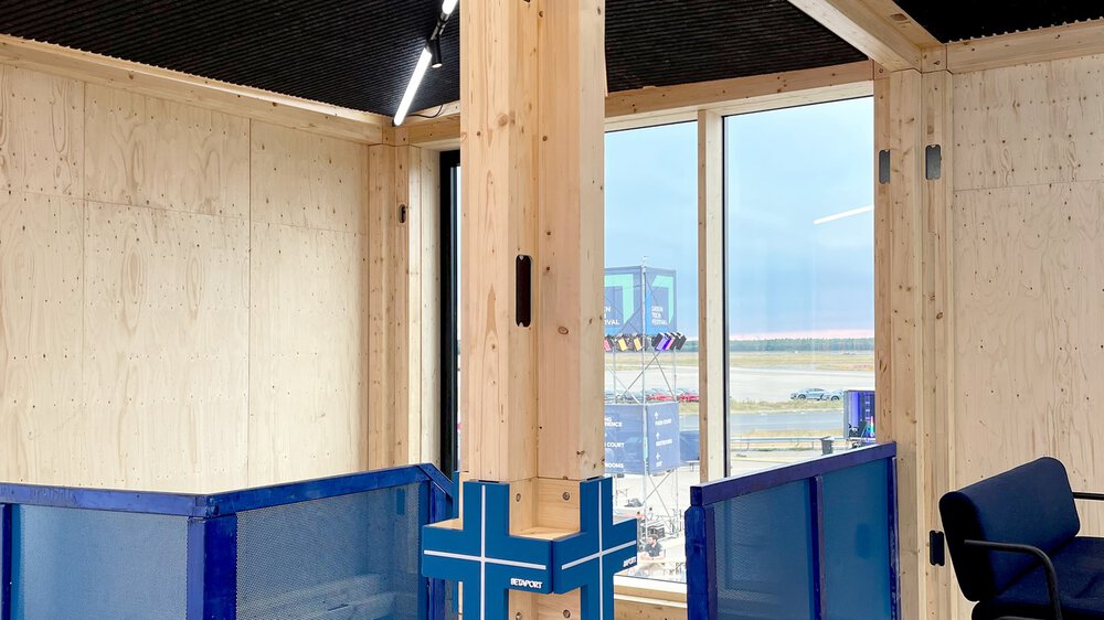 Ein heller Raum mit einer Holzsäule und kreuzförmigen Deckenverstrebungen vor einem Treppenabgang mit blauen Geländern.
