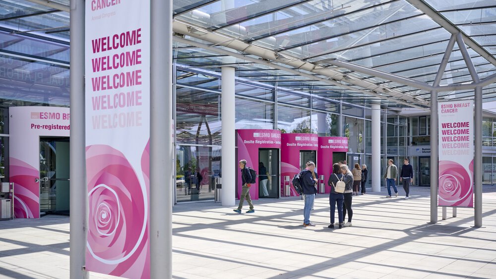 People enter a modern glass building for an ESMO breast cancer event, where large welcome banners are displayed.