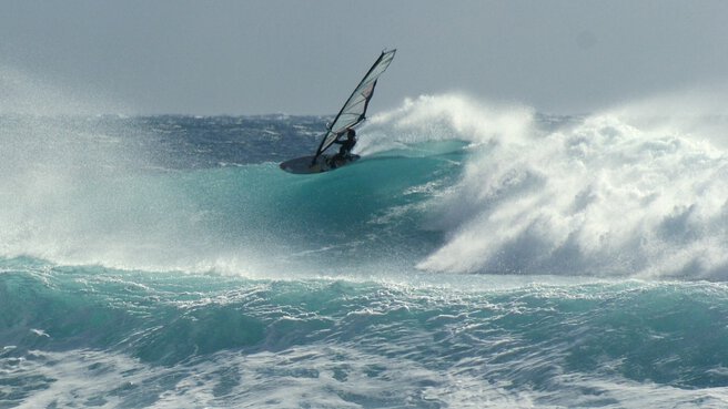 Ein Windsurfer reitet auf einer großen Meereswelle bei bewölktem Himmel.