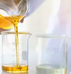 A person pours a yellow liquid from a beaker into a measuring cylinder, with laboratory equipment in the background.