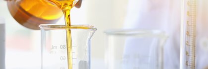 Eine Person gießt eine gelbe Flüssigkeit aus einem Becherglas in einen Messzylinder, mit Laborgeräten im Hintergrund.