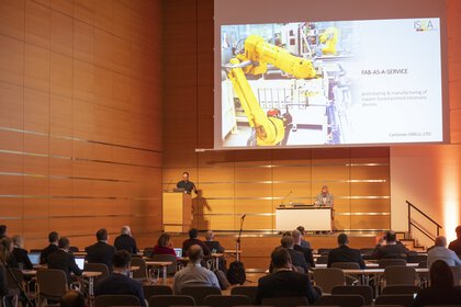 Menschen nehmen in einem Konferenzraum an einer Präsentation teil, auf einer großen Leinwand ist ein Roboterarm zu sehen.