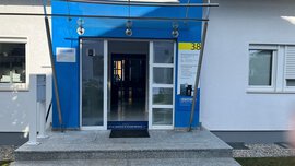 Eingang zu einem blau-weißen Gebäude mit Glastüren, Stufen und der Nummer 38 auf einem gelben Schild.