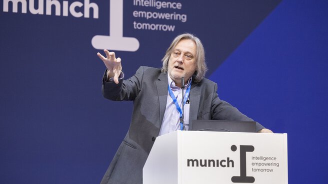 A speaker with gray hair gives a presentation on stage, gesturing with his hand as he speaks. He stands behind a podium featuring the logo “munich I – intelligence empowering tomorrow,” set against a dark blue backdrop.