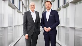 Zwei Männer in einem schwarzen und einem blauen Anzug stehen in einem mit Fenstern verglasten Gang und lächeln.