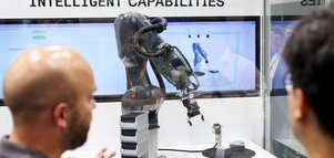 Two men are standing in front of a glass display case containing a black robotic arm. The case is labelled ‘INTELLIGENT CAPABILITIES’.