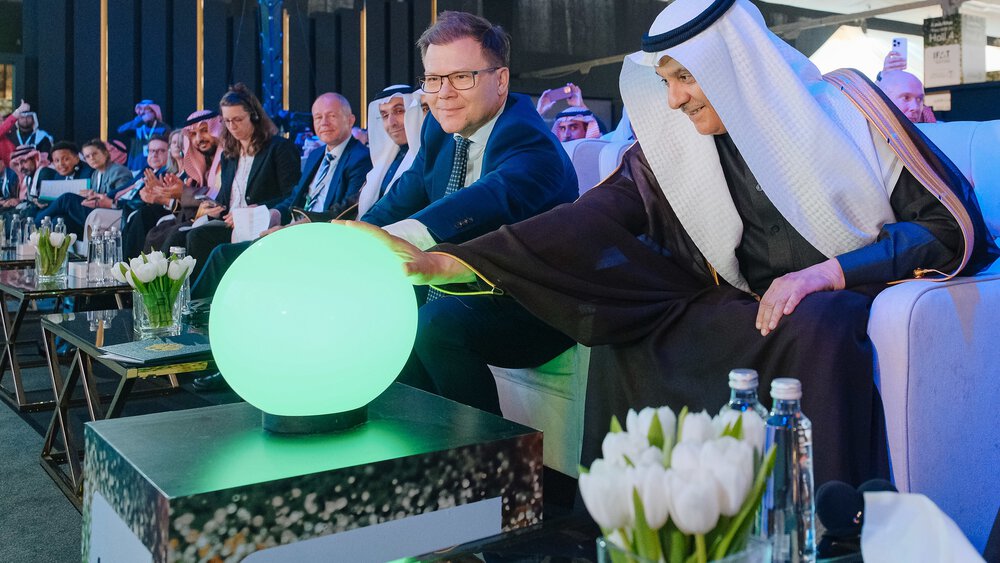 Two men are sitting on armchairs, each holding one hand on a green glowing ball standing on a small table.