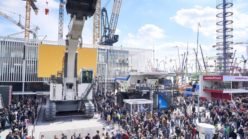 A large crowd of people at a construction machinery exhibition inspect huge machines and cranes under a partly cloudy sky.